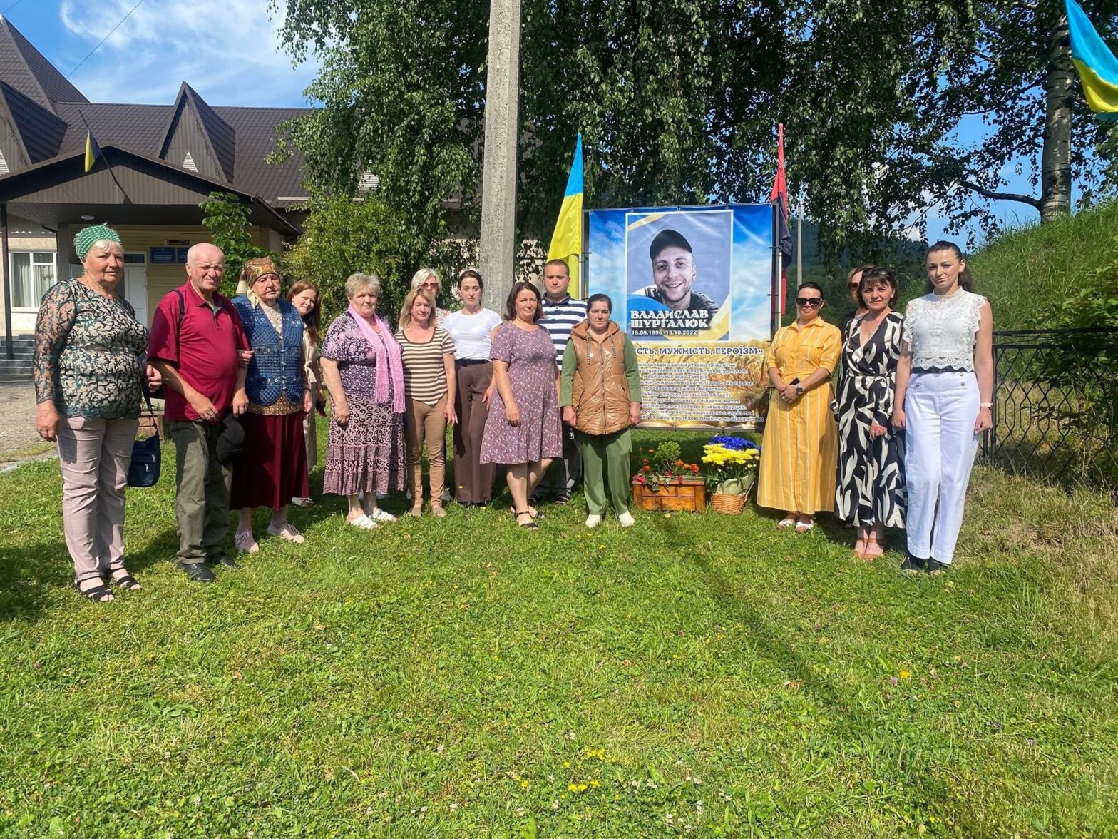 Група осіб стоїть на траві навколо меморіалу загиблому військовому. Коло меморіалу стоять жовто-сині квіти