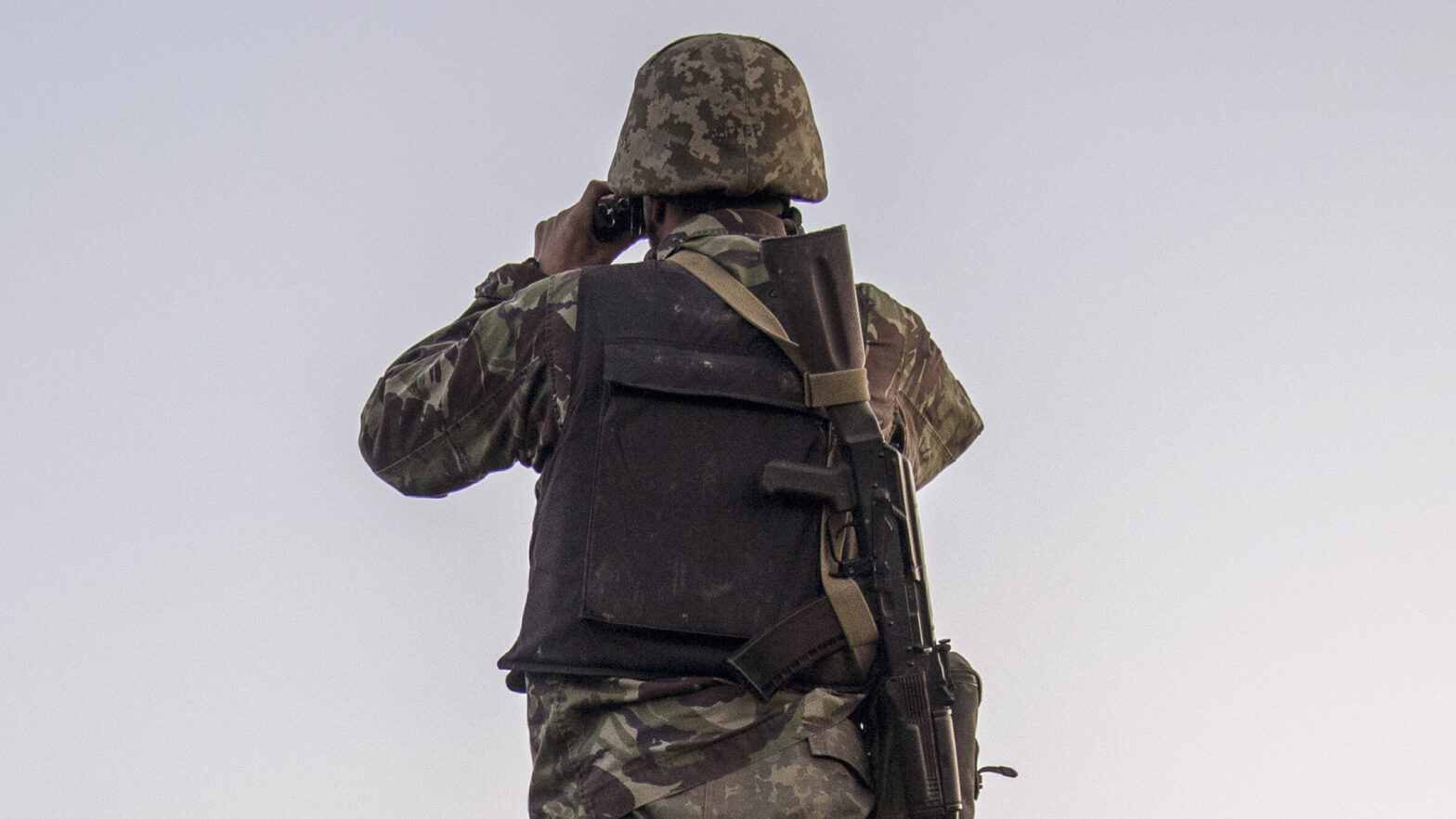 Чоловік у військовій формі та касці стоїть спиною та дивитися з бінокля в далечінь. Обличчя не видно, на фоні - небо