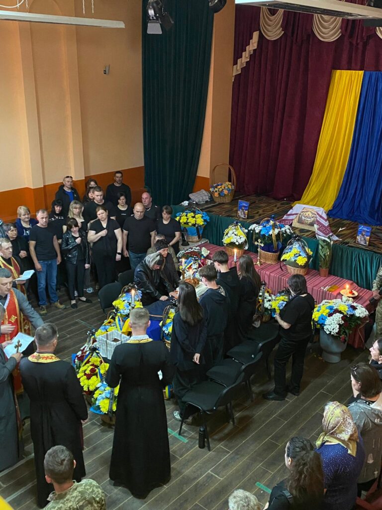 Натовп людей у приміщенні, труна посередині. Збоку на сцені видніється великий прапор України