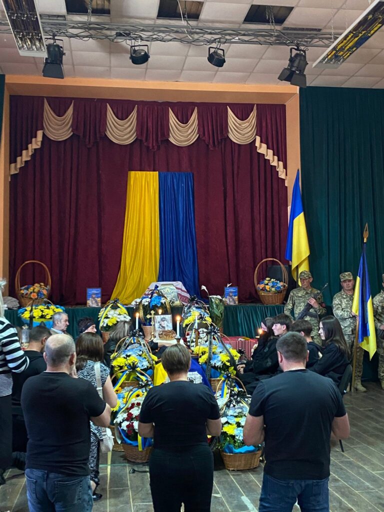У приміщенні троє осіб на колінах моляться перед труною, на фоні - прапор України. Біля труни сидять люди
