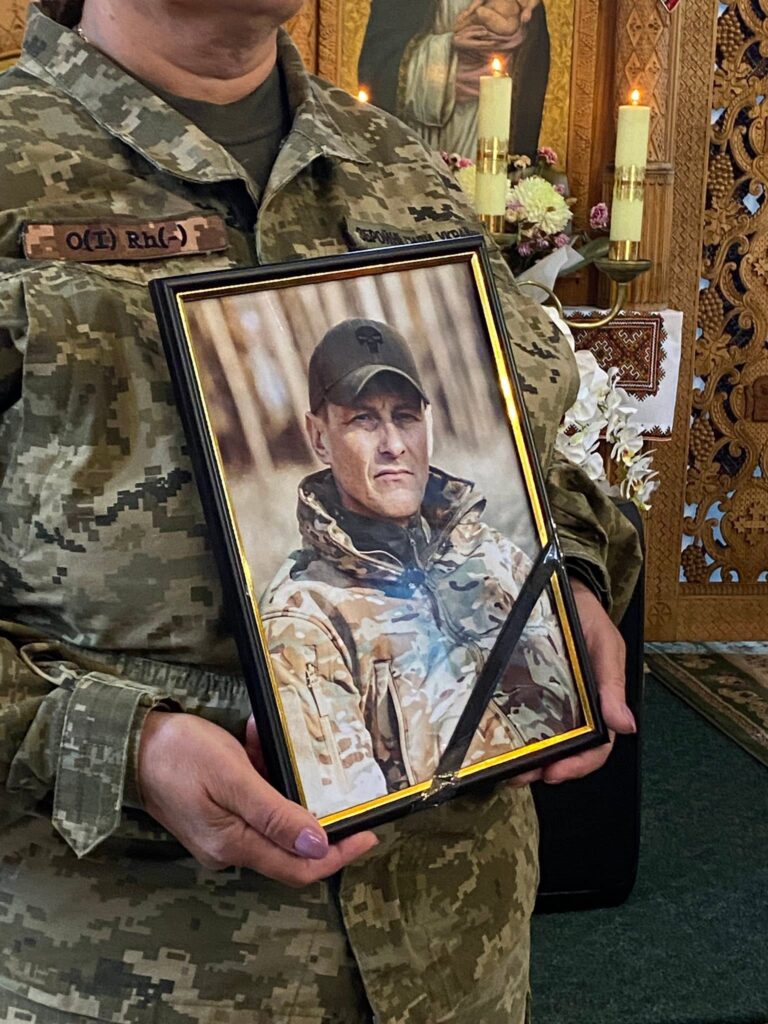 Людина у військовій формі тримає світлину в чорній рамці та чорною стрічкою. На світлині чоловік у віці 40+ років дивиться, трохи прищурившись, у військовій формі та кепкою на голові.