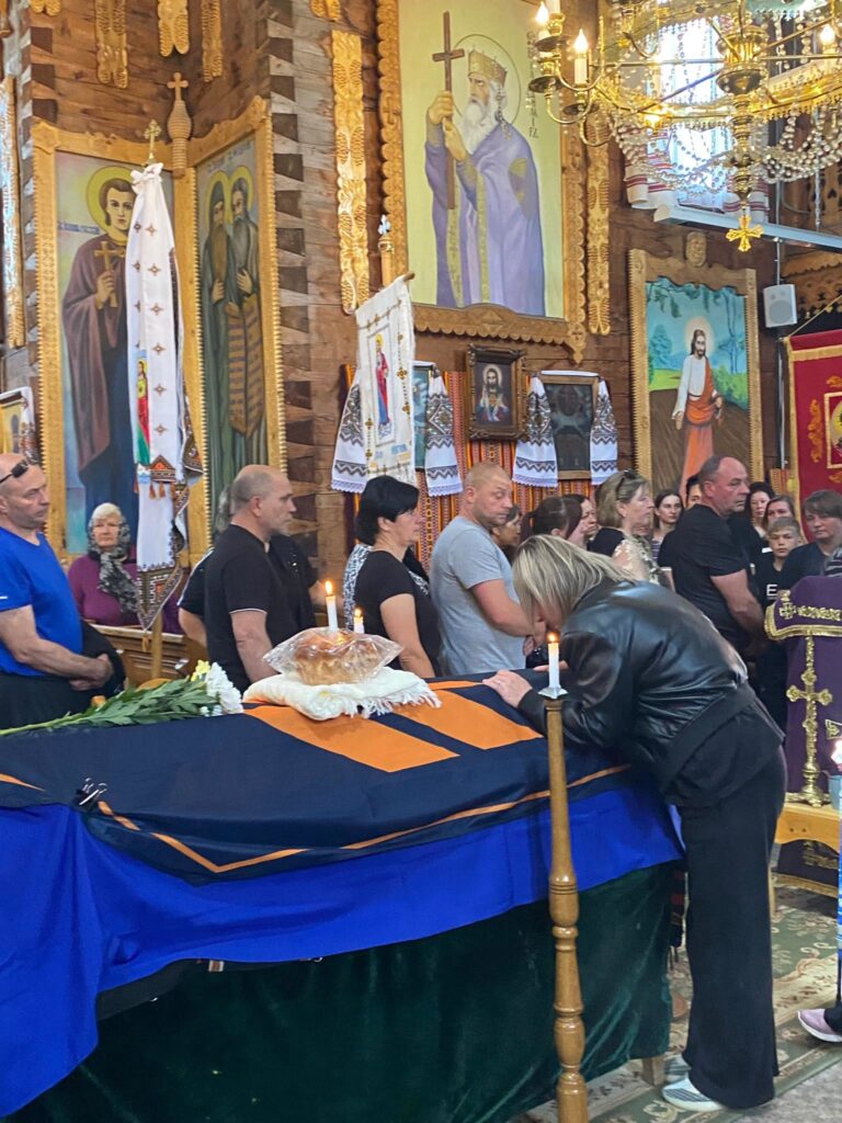 Жінка у чорній одежі схилилася до труни чолом. Труна вкрита українським прапором та прапором 3-ї ОШБр, стоїть у церкві. Навколо нього підсвічники та ікони, стоять люди.