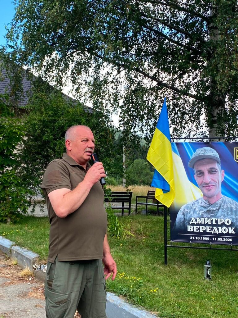 Чоловік у футболці та штанях кольору хакі щось говорить у мікрофон, позаду видно частину банеру зі світлинами полеглих
