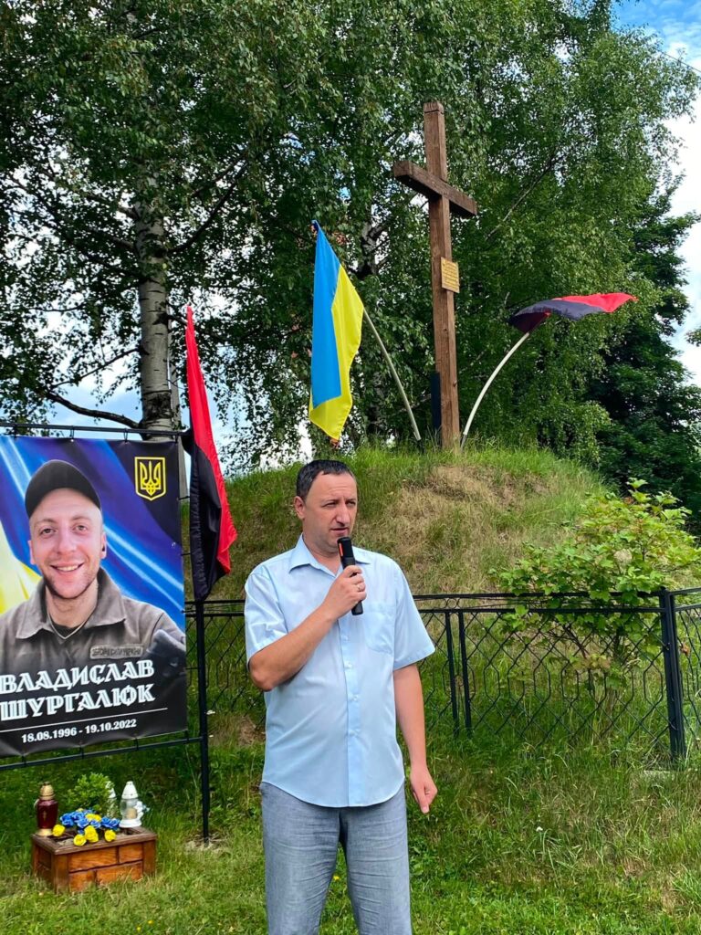 Чоловік з мікрофоном у блакитній сорочці з коротким рукавом та штанах синього відтінку, позаду видніється пам'ятний банер з обличчям юнака