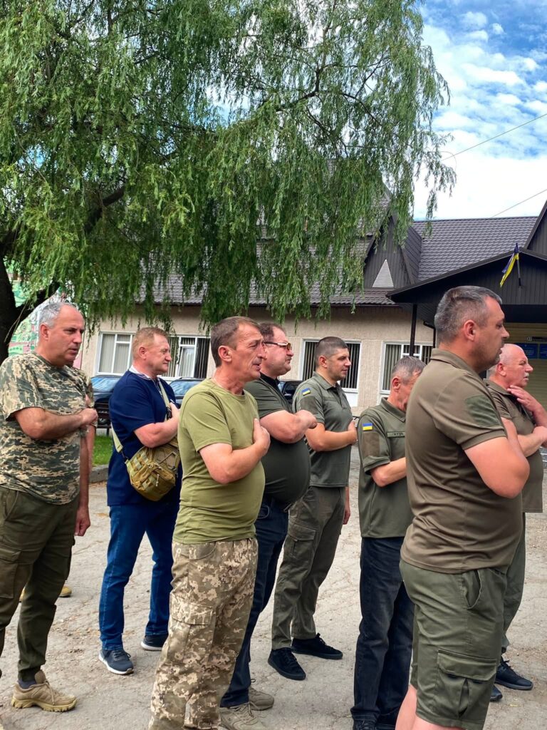 Декілька чоловіків у футболках кольору хакі та штанях з пікселями стоять, поклавши руку на серце