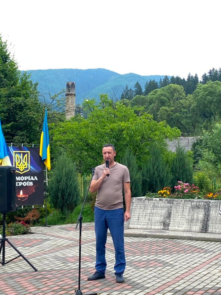 Чоловік з мікрофоном стоїть на вимощеній площі, трохи збоку видніється меморіальний банер та український прапор