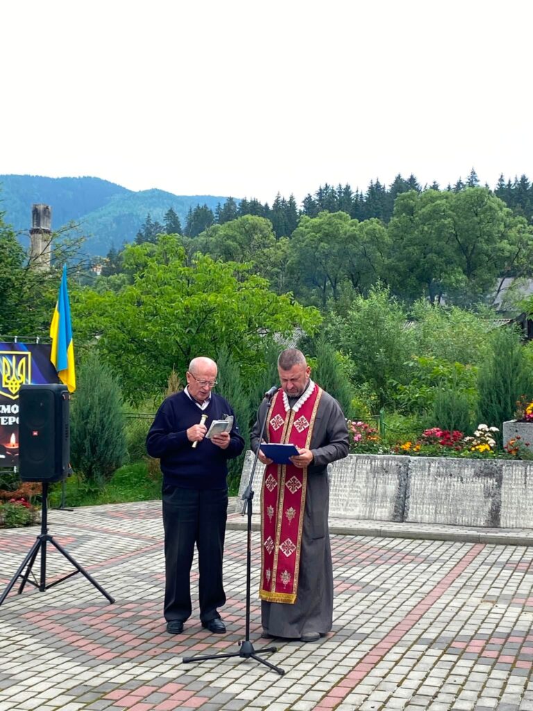 Два чоловіки стоять біля стійки з мікрофоном, один з них в церковній рясі та схилив голову над книгою