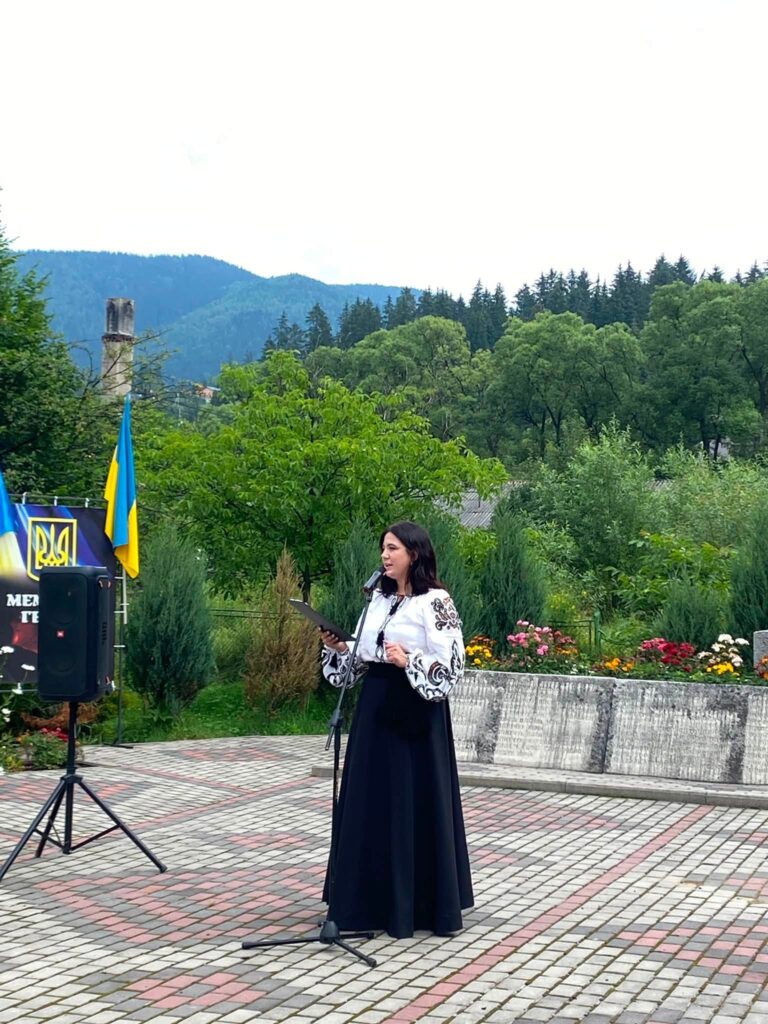 Жінка у вишиванці та чорній довгій спідниці з планшеткою стоїться біля стійки з мікрофоном на вимощеній площі