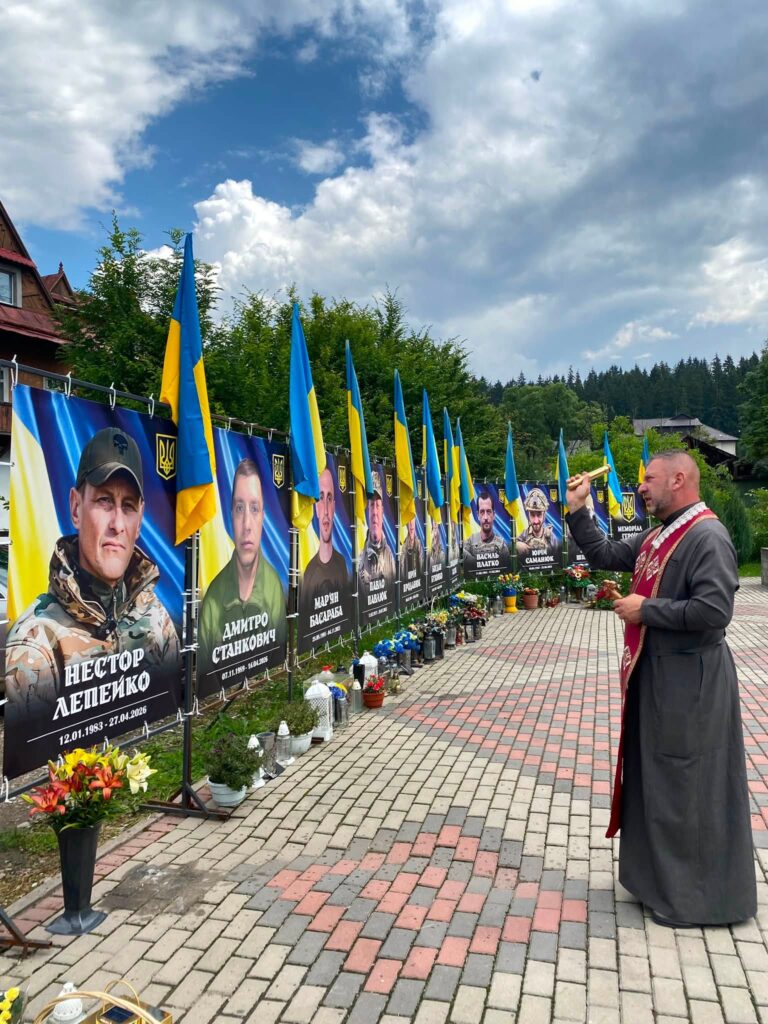 Чоловік у церковній рясі змахує кропилом перед меморіальним банером, де зображений чоловік у військовій формі