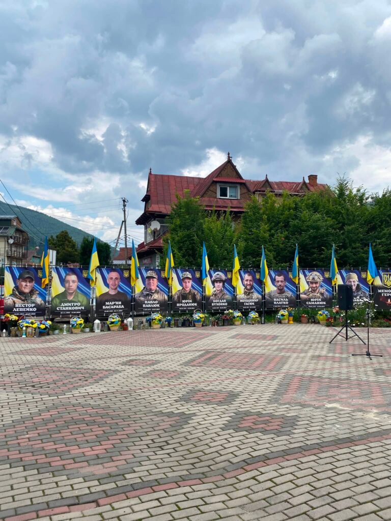 Ряд меморіальний банерів з обличчями чоловіків, поряд з кожним банером стоять синьо-жовті стяги