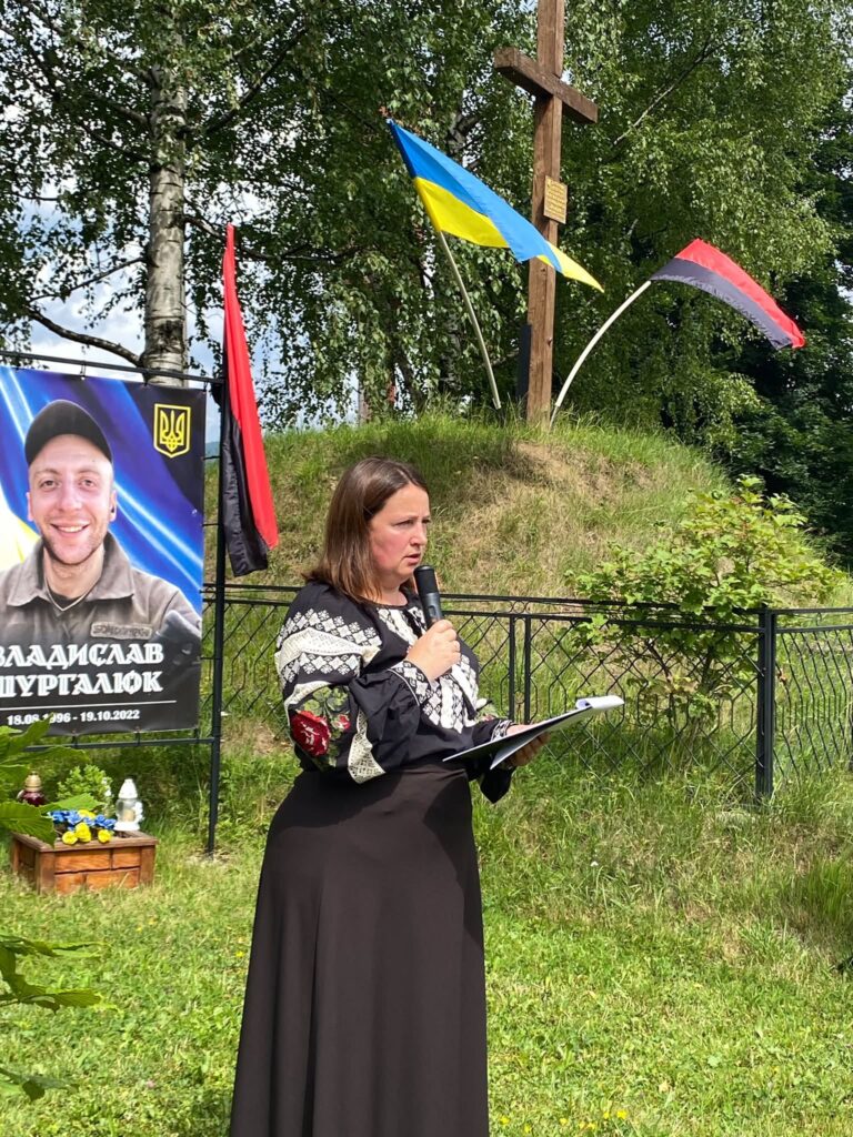Жінка у чорній вишиванці та довгій чорній спідниці стоїть з мікрофоном та аркушами, позаду видніється світлина чоловіка та прапори