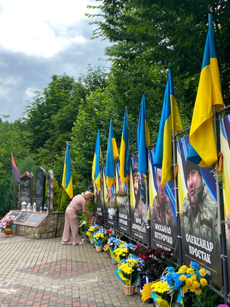 Жінка ставить кошик жовто-блакитних квітів до меморіального банера