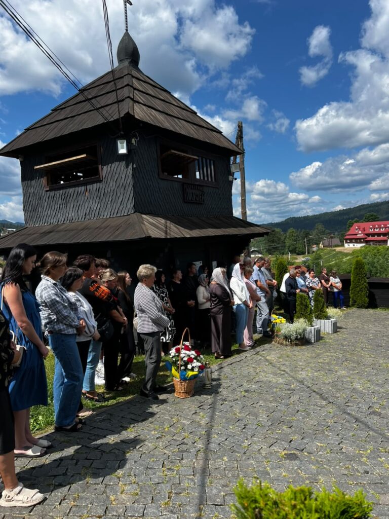 Невеликий натовп людей стоїться на вимощеному подвір'ї, позаду них чорна дерев'яна дзвіниця.