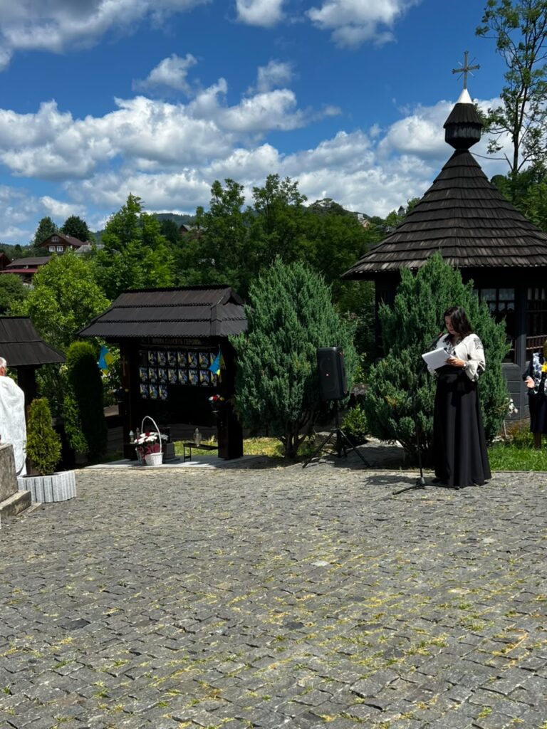 На вимощеному подвір'ї стоїть жінка з чорним волоссям у чорній спідниці та вишитій сорочці біля стійки мікрофона та стенду з полеглими. На фоні зелені дерева та дерев'яна стріха з хрестом
