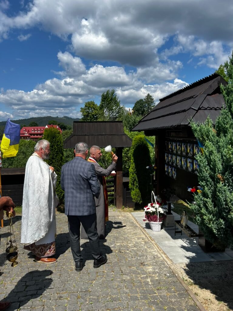 Троє чоловіків стоять навпроти стенду на честь полеглих, один тримає у руці кропило, другий у білій рясі стоїть трохи позаду, третій у костюмі стоїть спиною