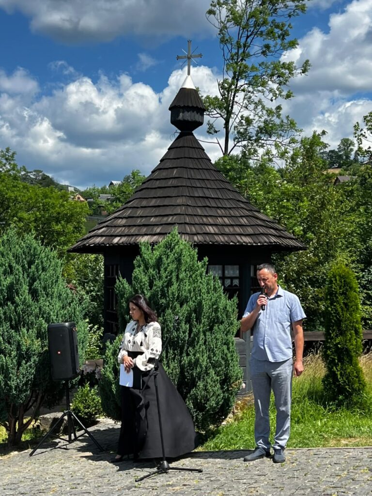 Двоє людей стоять на фоні зелені та чорної дерев'яної споруди, чоловік тримає у руках мікрофон, друга жінка у чорній спідниці та вишитій сорочці тримає в руках листи паперу
