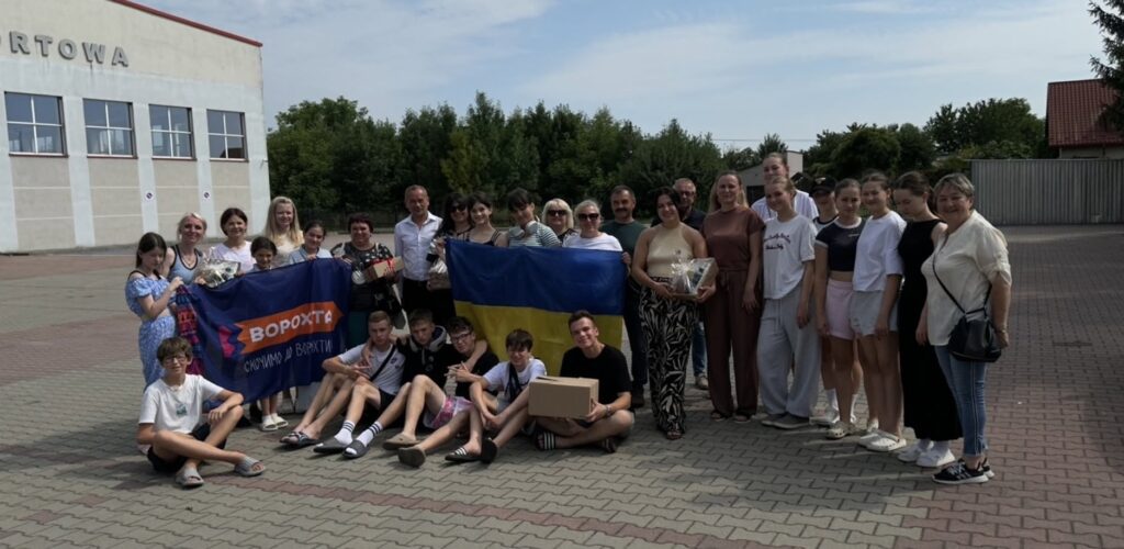 Діти і дорослі стоять напівколом перед камерою, тримаючи у руках жовто-синій прапор та фіолетовий прапор, де на помаранчевому фоні пише ВОРОХТА