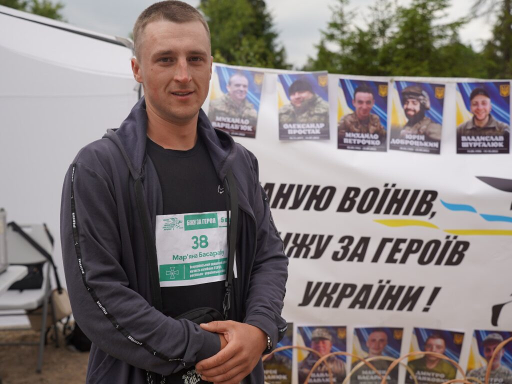 Чоловік середнього віку стоїть перед камерою, позаду нього видніється банер з текстом "Шаную воїнів, біжу за Героїв України" та фотографіями чоловіків у військові формі